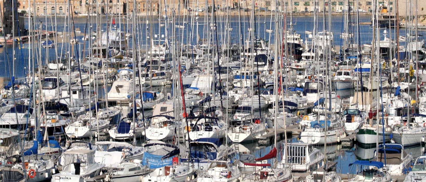 Rows of moored sailing yachts in a harbour marina, with honey-coloured stone buildings rising on the hill behind.