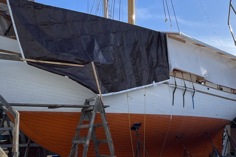 A wooden hull ashore on stands in a boatyard, covered over amidships.
