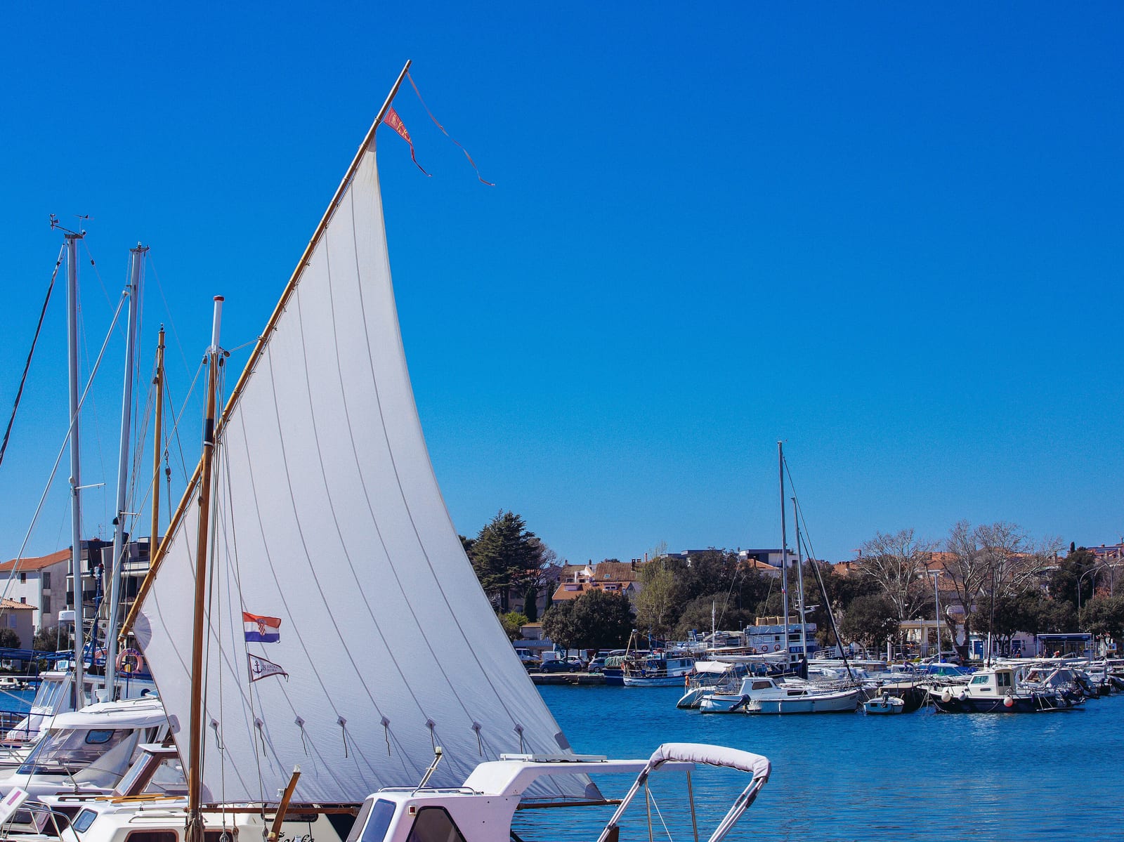 A white mainsail raised on a yacht under way, with moored boats and a wooded shoreline behind.