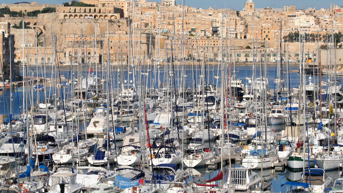A marina basin packed with moored sailing yachts below a stone harbour town.