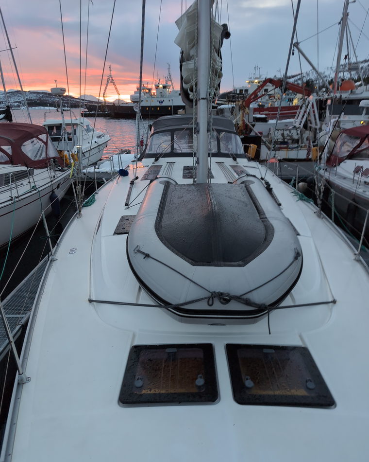 A yacht foredeck with a stowed liferaft and deck hatches at dusk in harbour.
