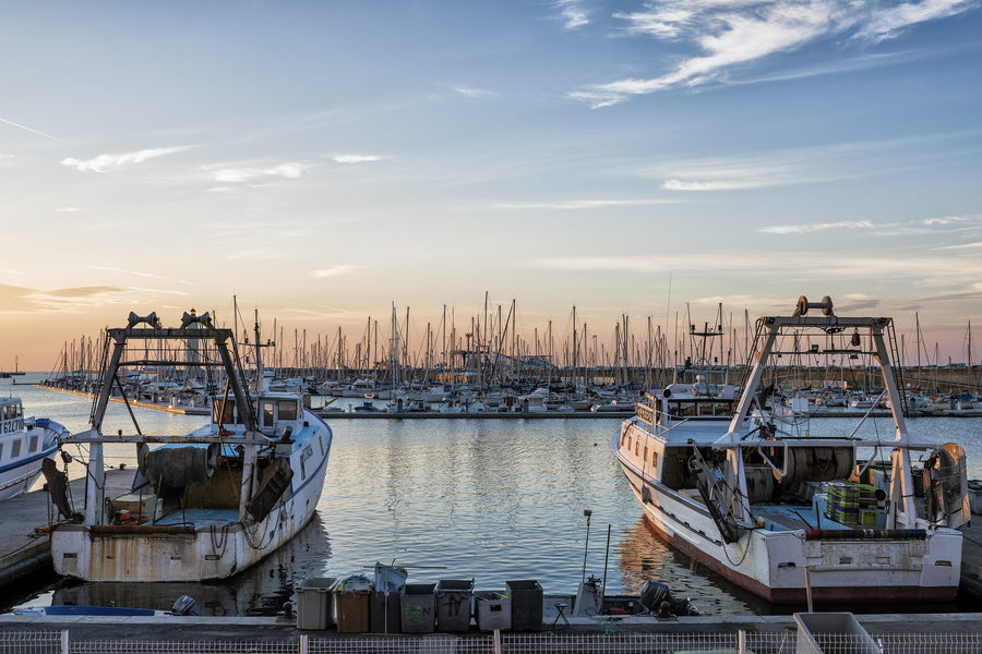 Two working boats moored along a harbour quay with a mast-filled marina behind.