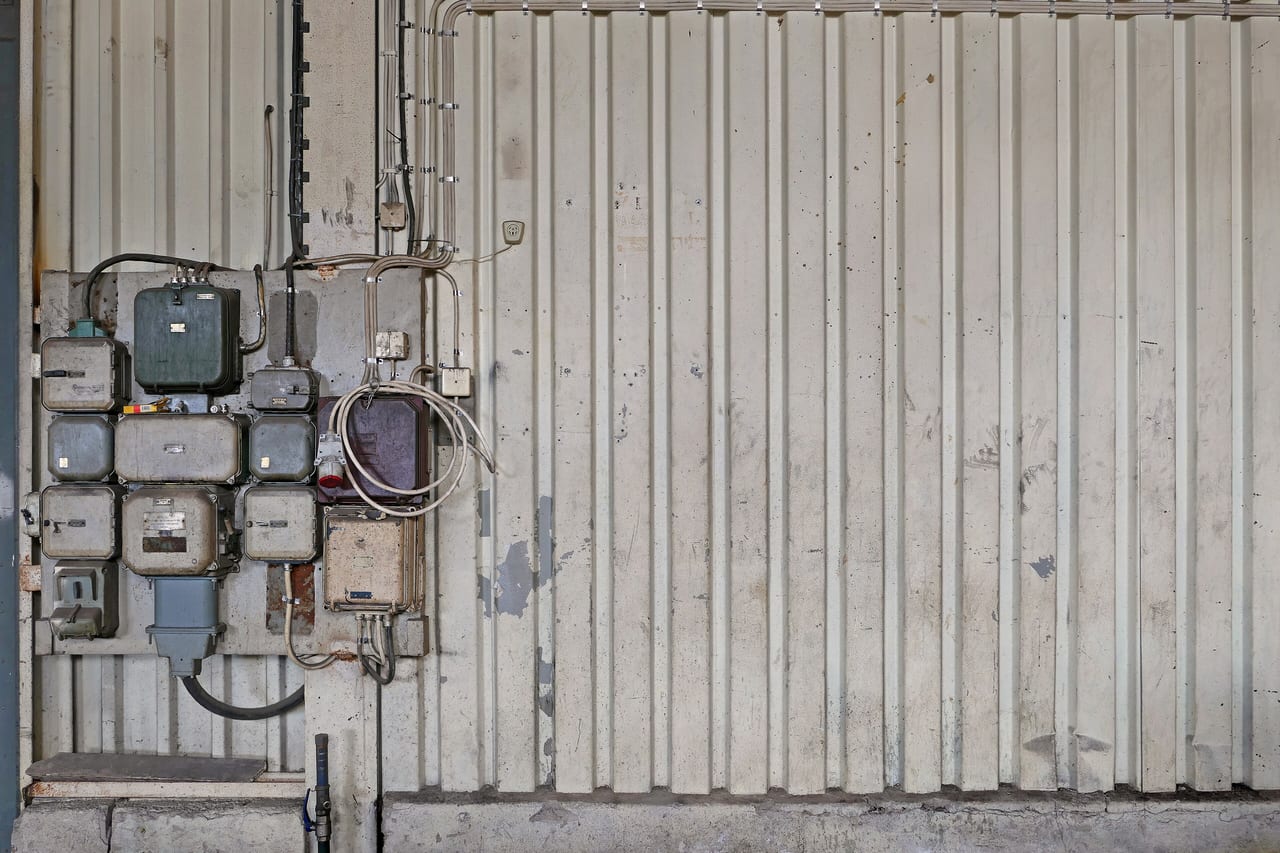 A wall-mounted distribution board carrying meters and metal enclosures, with conduit risers running up the workshop wall beside it