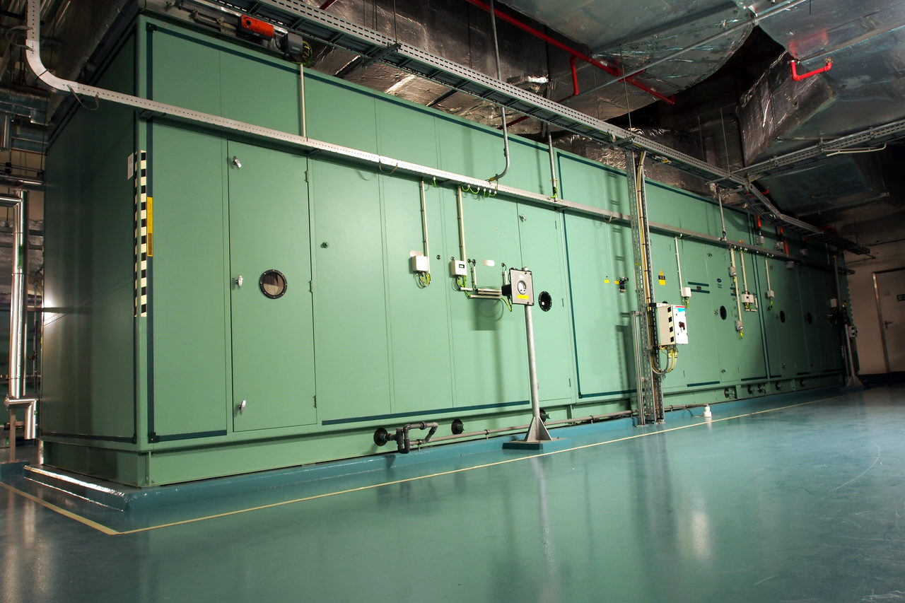 A long pale green air handling unit running down a plant room, with insulated ductwork above it and drain pipework at its base
