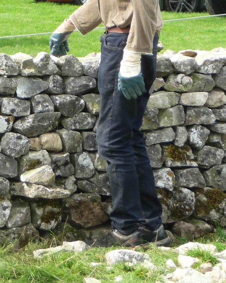 A dry stone wall of grey fieldstone part-built, with a person in work gloves standing behind it, cropped at the chest so no face is visible.