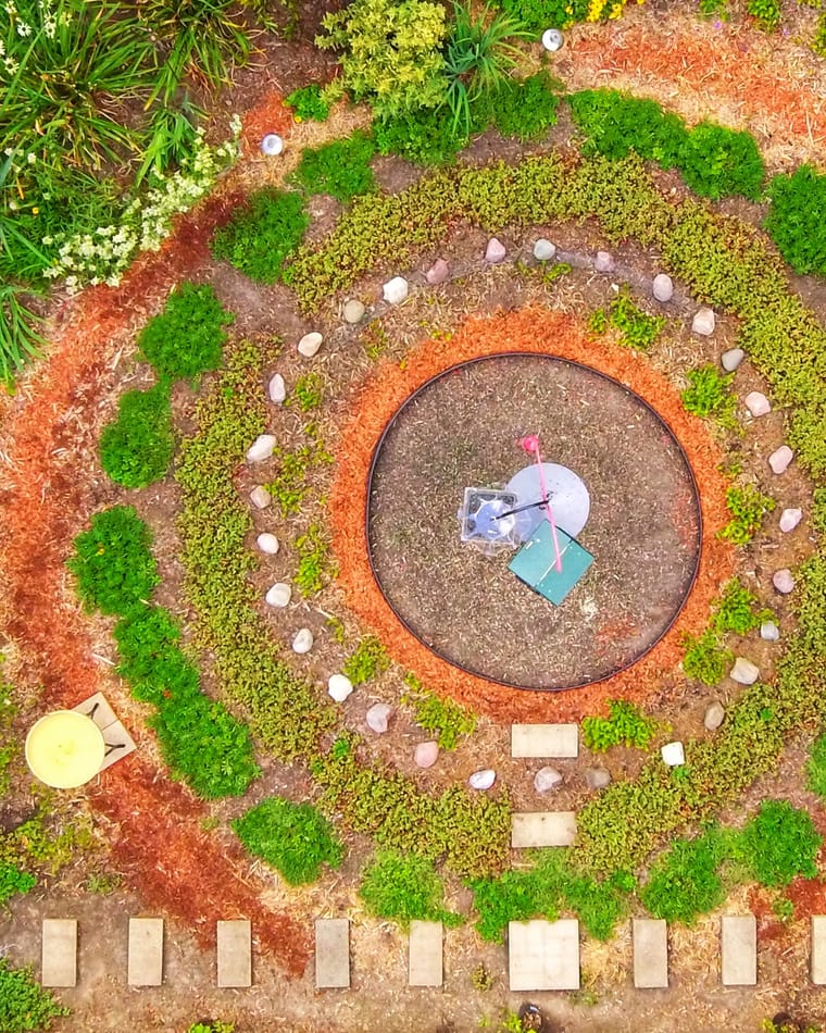 A circular garden bed seen from directly above, with rings of young plants and a ring of pale stones around a central gravel circle.
