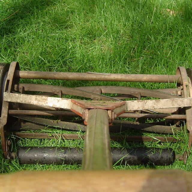 A close view at ground level of an old push reel mower, its blades and roller resting on half-cut grass.