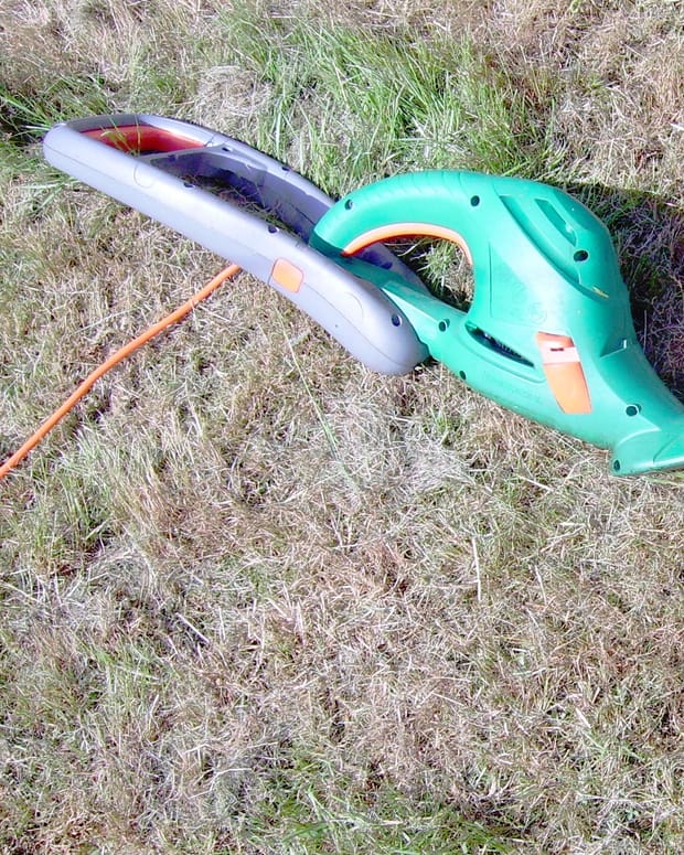A green electric hedge trimmer lying on dry grass with its orange cable trailing behind it.