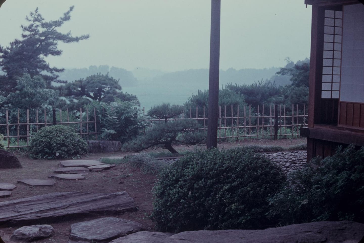 A stone path beside clipped evergreen planting, seen from the shade of a timber veranda with a valley beyond.