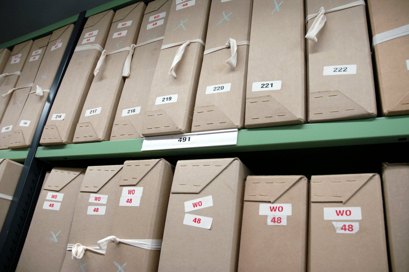 Labelled archive boxes packed along two levels of green metal repository shelving, each box tagged with a reference card.