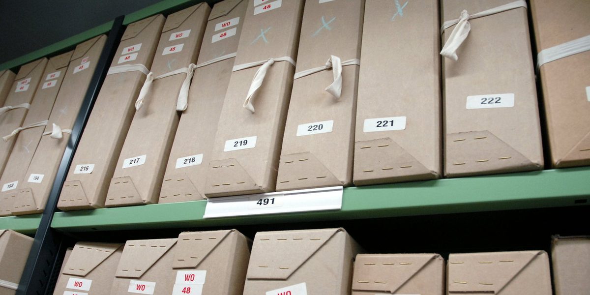 A run of numbered archive boxes standing on green repository shelving, their reference labels reading 216 to 222 in sequence.
