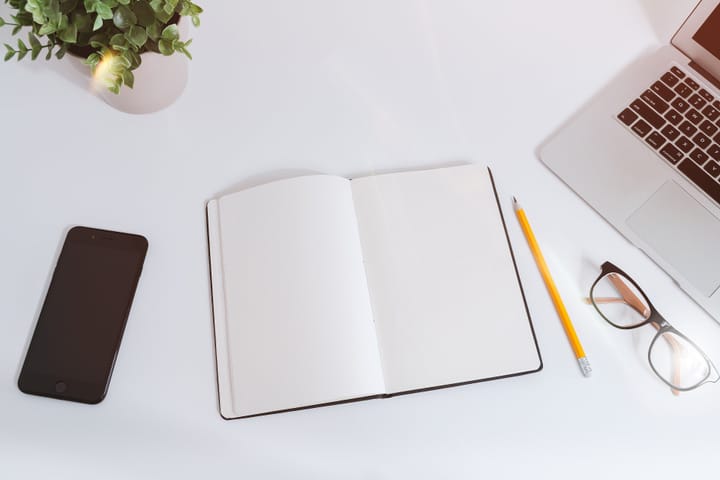A white work table seen from above, with a blank notebook, a yellow pencil, glasses, a smartphone and the corner of a laptop.