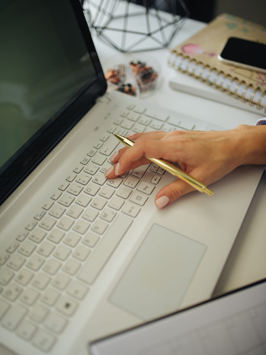 A hand holding a gold pen over a silver laptop, beside a notebook and a phone.