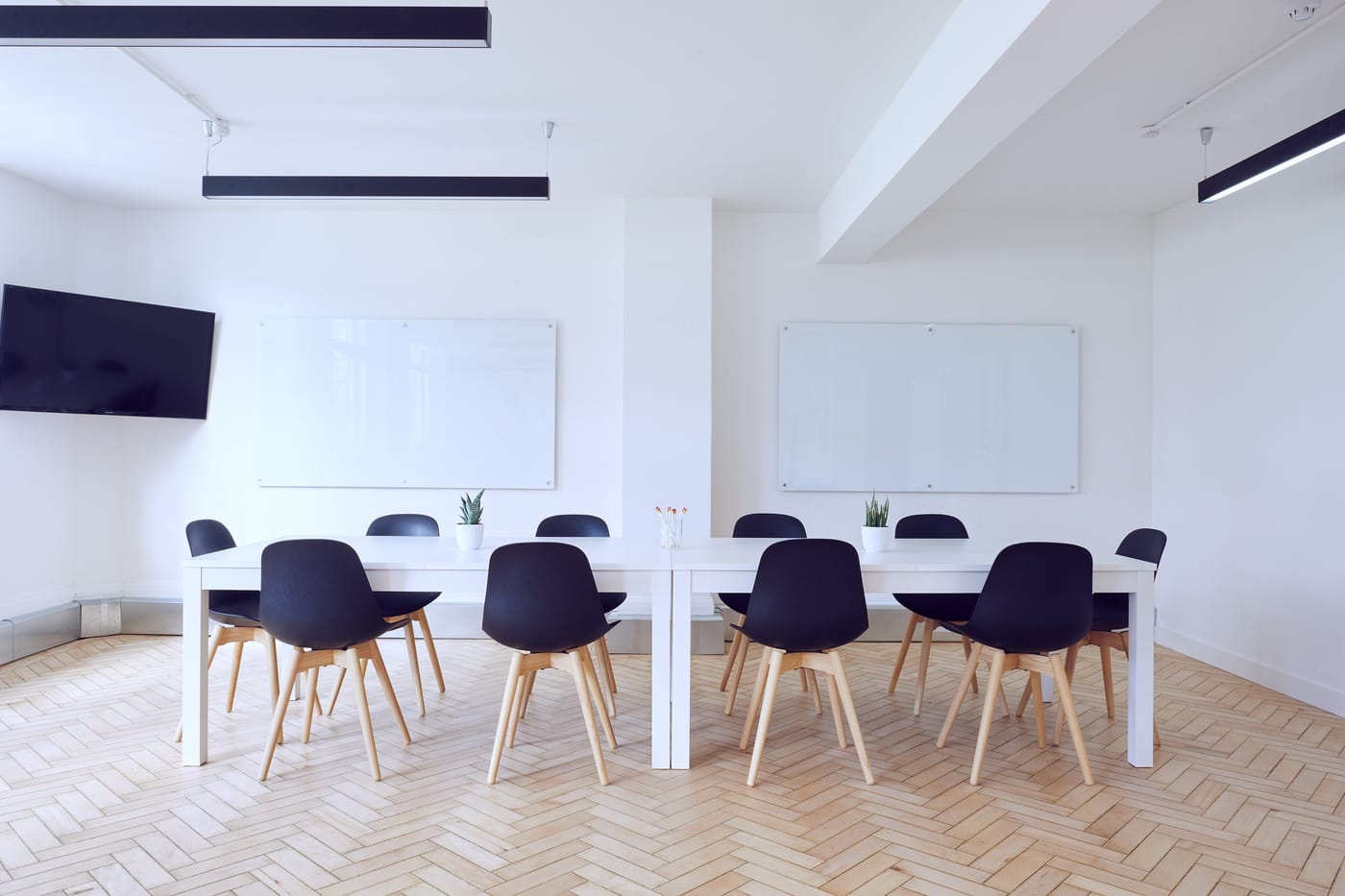 A bright meeting room with white tables, navy chairs, whiteboards and a wall screen above a herringbone floor.