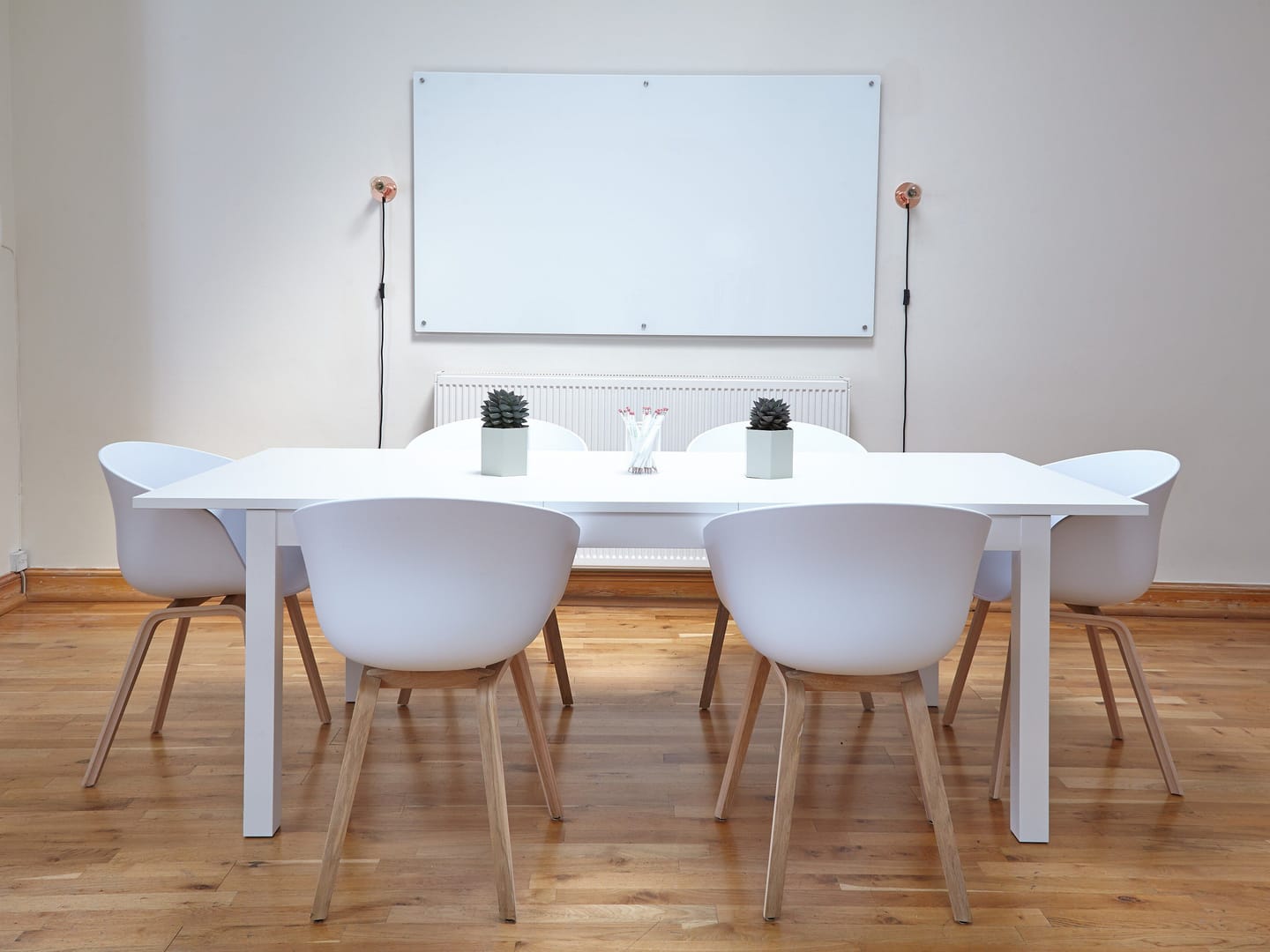 A bright meeting room with a long white table, white chairs, a blank whiteboard and potted plants on a wooden floor.