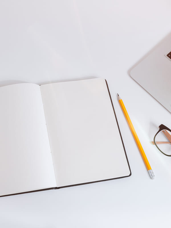 An open notebook, pencil, glasses and a laptop on a white desk