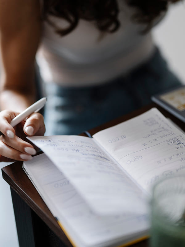 A hand with a pen working down a page of handwritten notes in an open notebook on a desk
