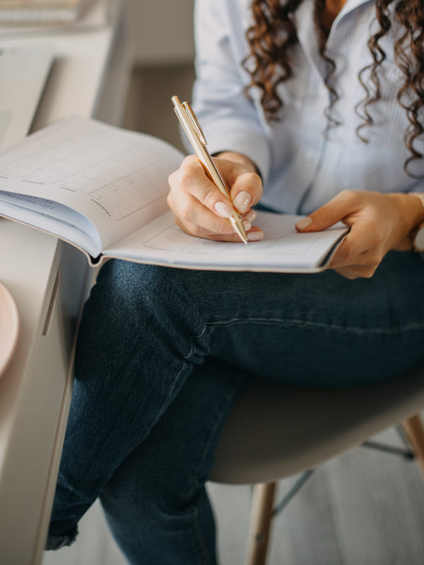 Someone writing notes into a ruled planner held on their lap at a desk