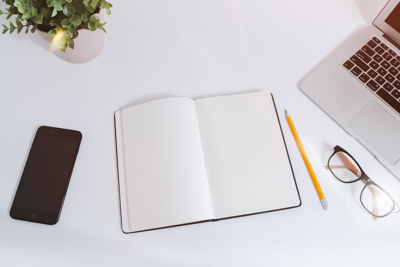 A white work table with handwritten notes, a smartphone and a laptop.