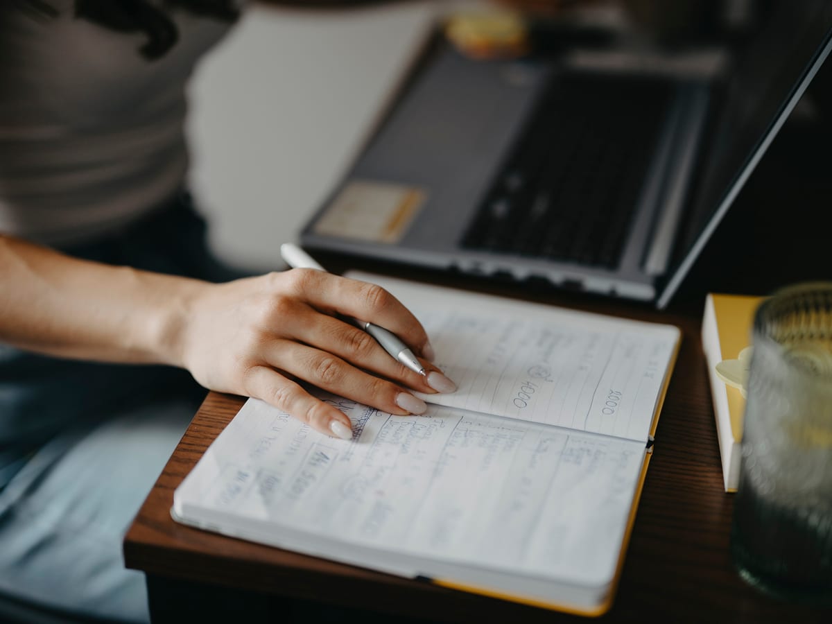 A person writing notes on paper beside an open laptop at a desk, face out of frame.
