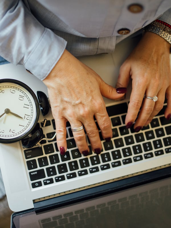Hands typing on a laptop with a small desk clock standing beside it