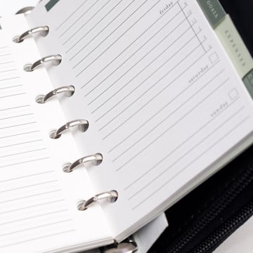 An open ring-bound planner showing blank weekday columns and goals and expenses dividers