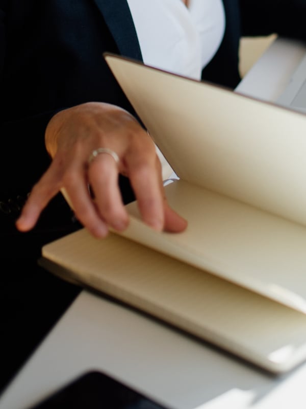 A hand turning to a page of an open notebook on a desk beside an open laptop