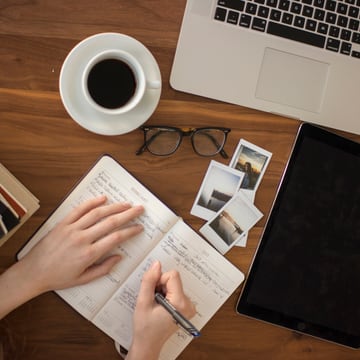 A desk with an open notebook being written in, a coffee, glasses and a tablet