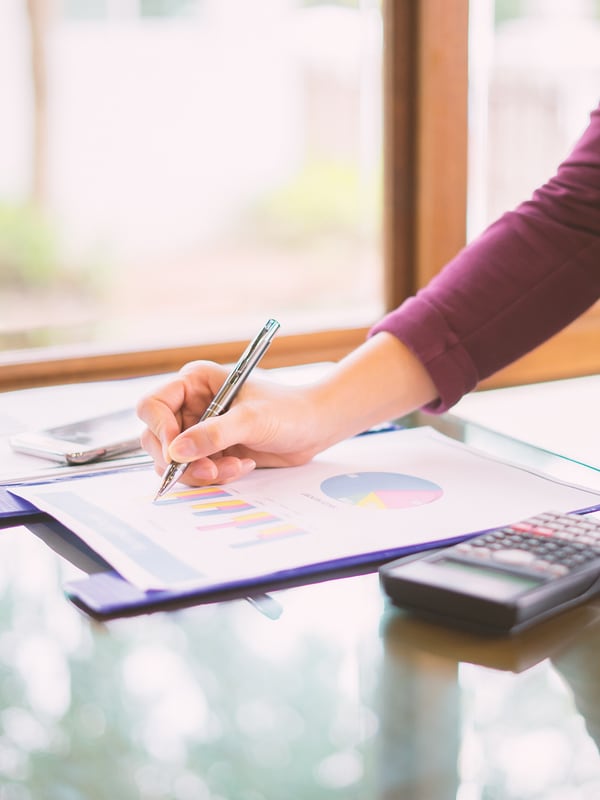 A hand marking a printed bar chart and pie chart with a pen, a calculator beside it