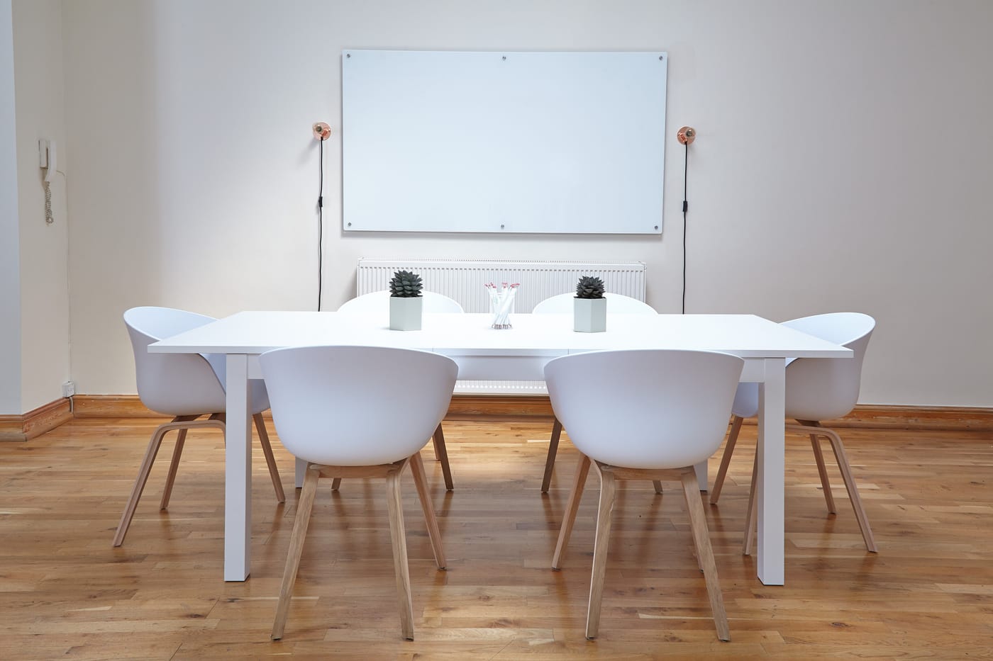 A small white meeting room with a white table, six white chairs and a blank whiteboard on the wall.