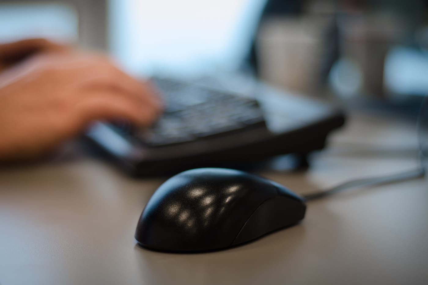 Two hands typing on a black keyboard, with a black mouse in focus in the foreground of a desk.