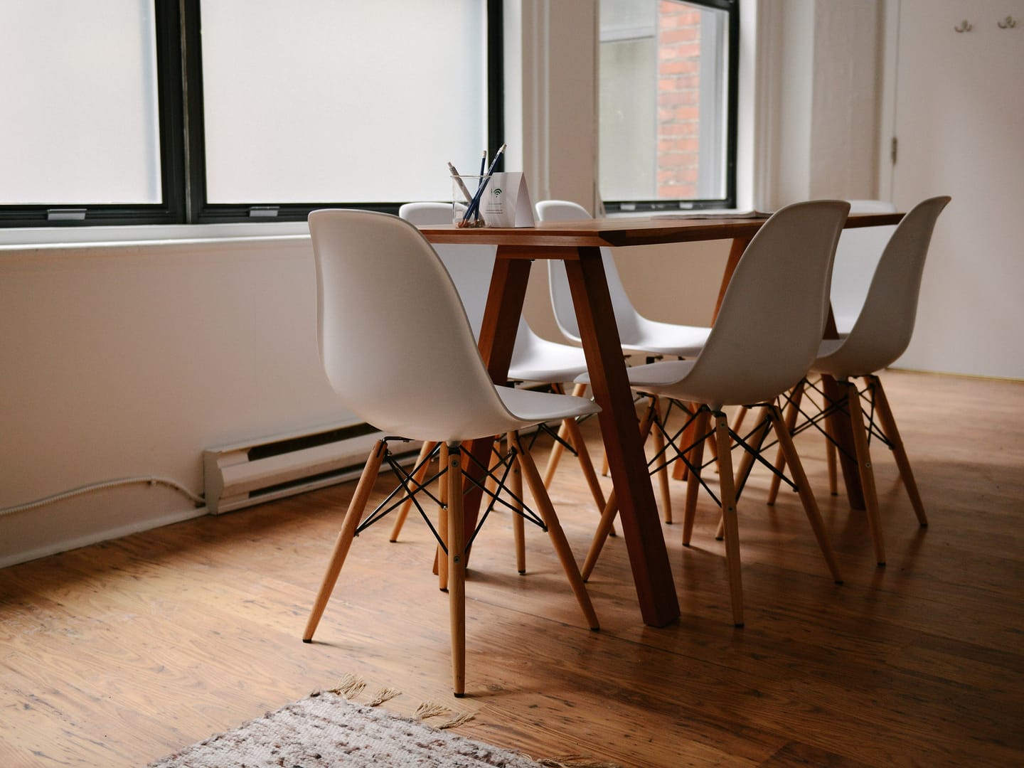 A wooden meeting table with white chairs in a quiet room lit by tall windows.