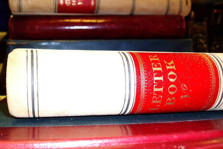The spine of a cream and red bound letter book, stacked between other volumes.
