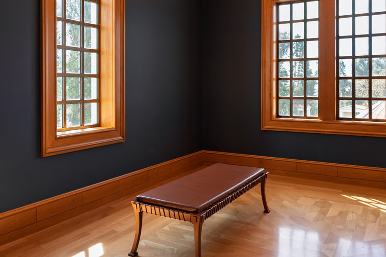 A bare room with dark painted walls, a wooden floor, two tall windows and one long bench.