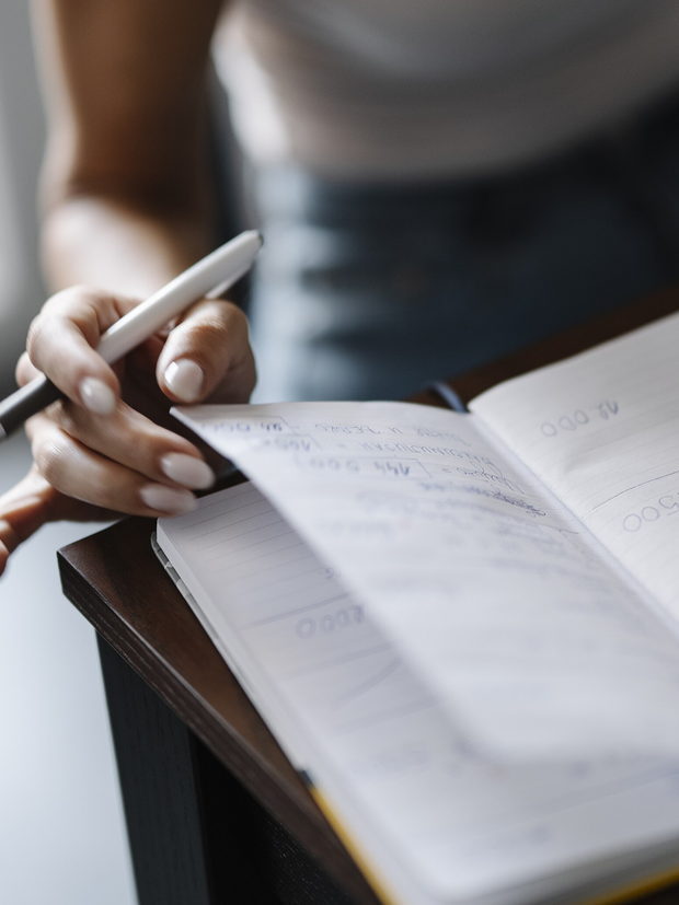 A hand resting a pen above an open notebook of handwritten figures, cropped short of any face.