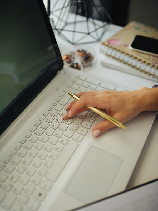 A hand resting on a laptop with a pen and a spiral notebook alongside