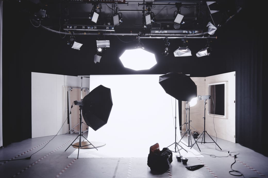 A darkened studio with a white cyclorama wall lit by overhead lamps and a softbox on a stand.