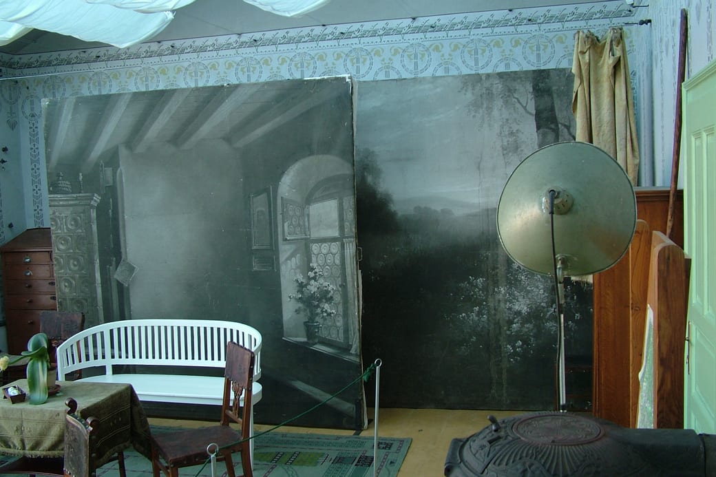 A portrait studio with a painted landscape backdrop, a white bench and a reflector lamp on a stand.