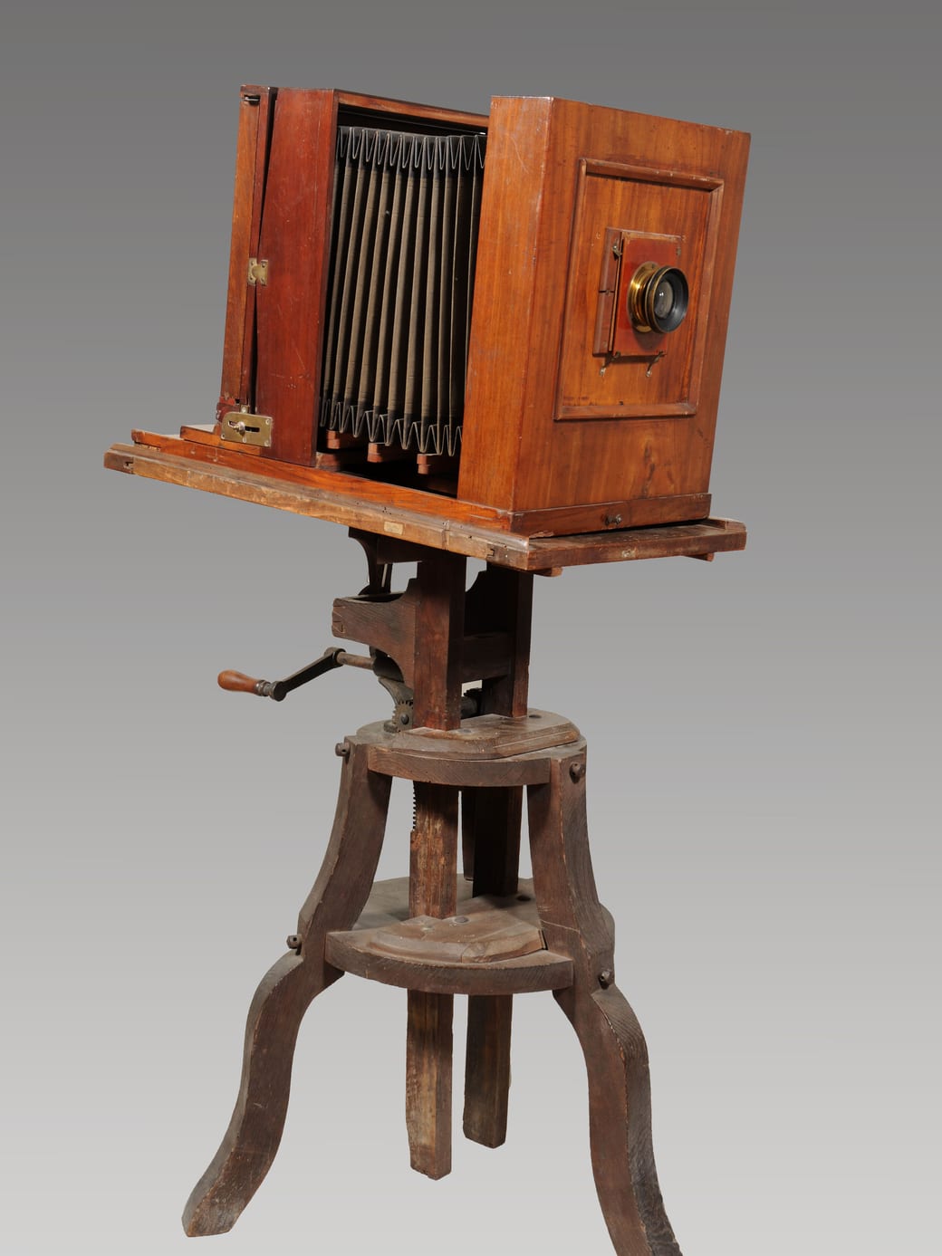 A wooden field camera mounted on its wooden tripod, photographed against a plain grey background.