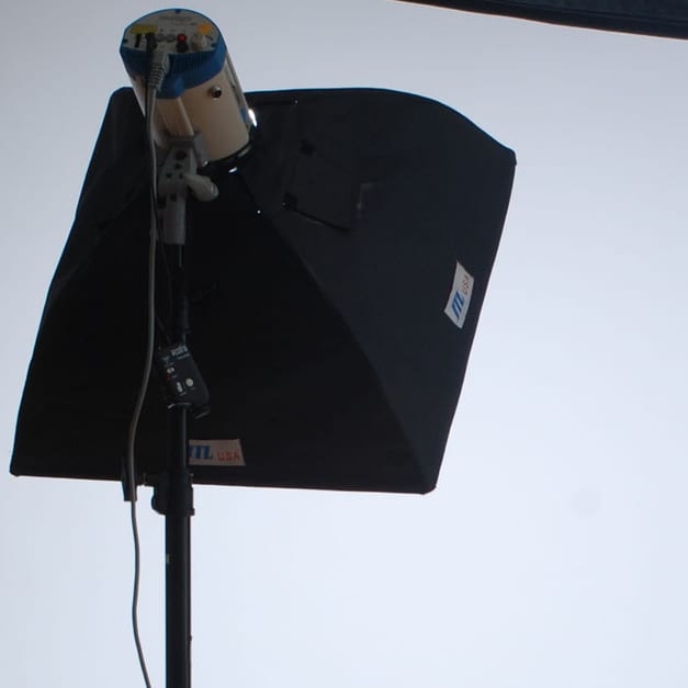 A black softbox with a strobe head mounted at its centre, carried on a stand in front of a plain white studio wall.