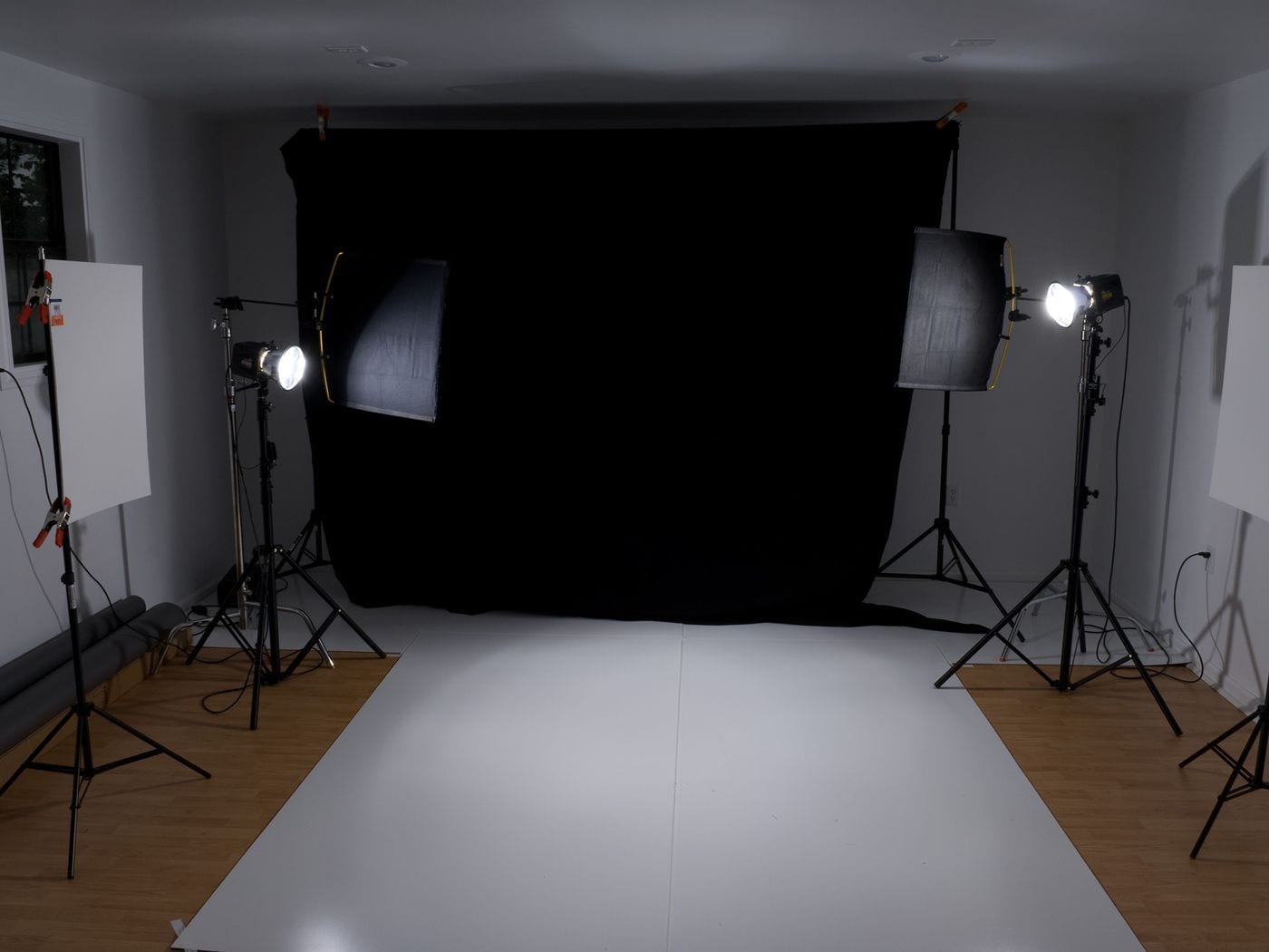 A black backdrop and a white floor between four lighting stands carrying softboxes and reflectors.