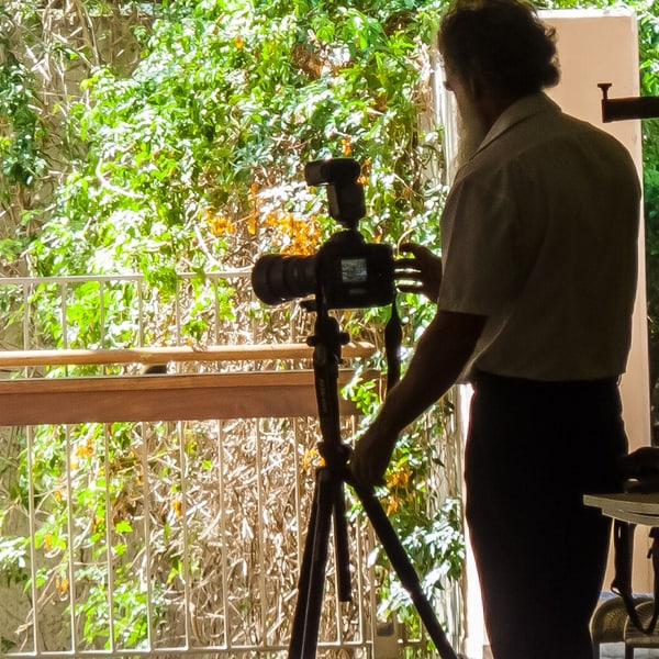 A photographer seen from behind at the camera on its tripod, one hand on the tripod leg, mid-sitting