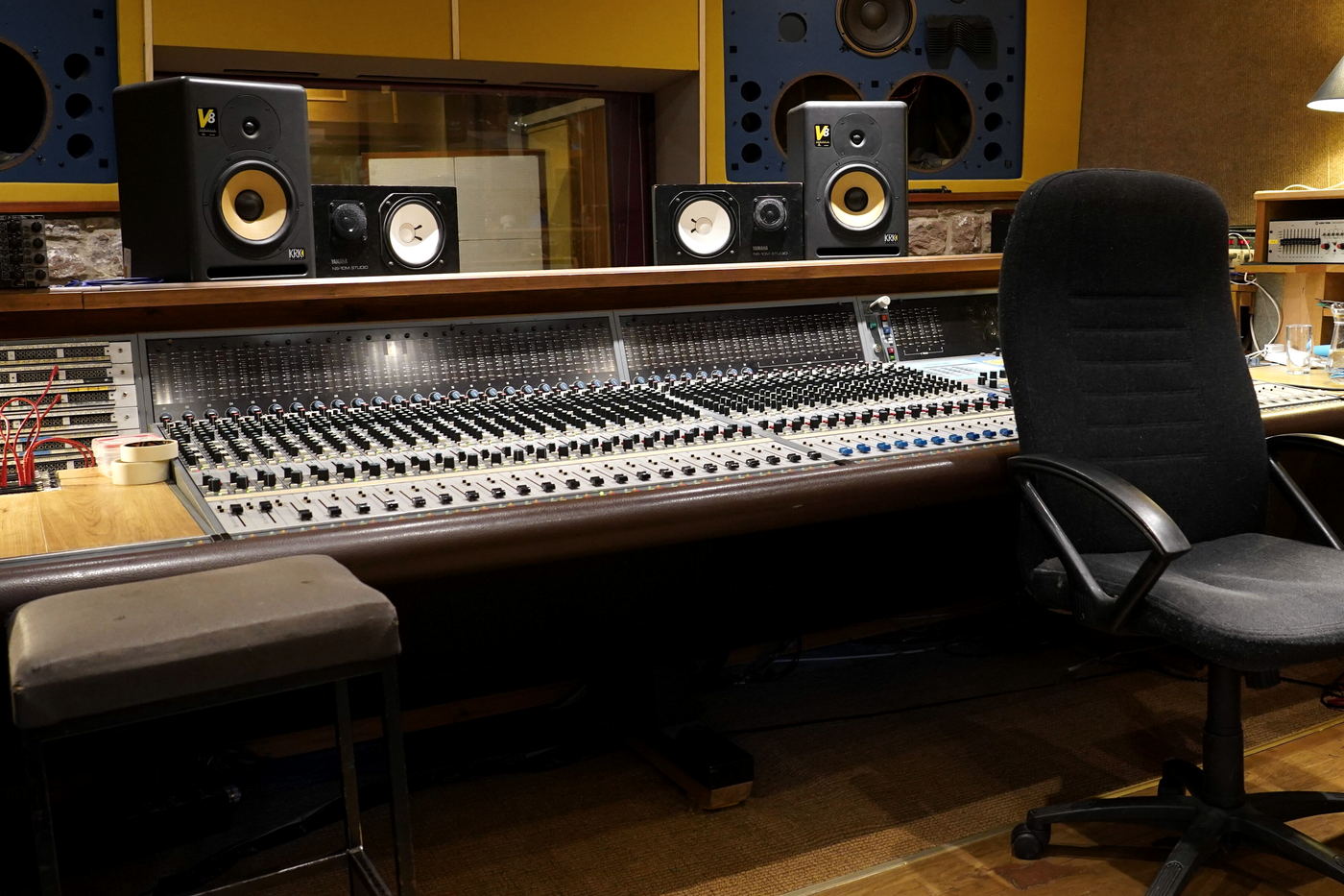 A long analogue console under a control-room window, with yellow-cone near-field monitors on the bridge and large soffit speakers set into a mustard wall.