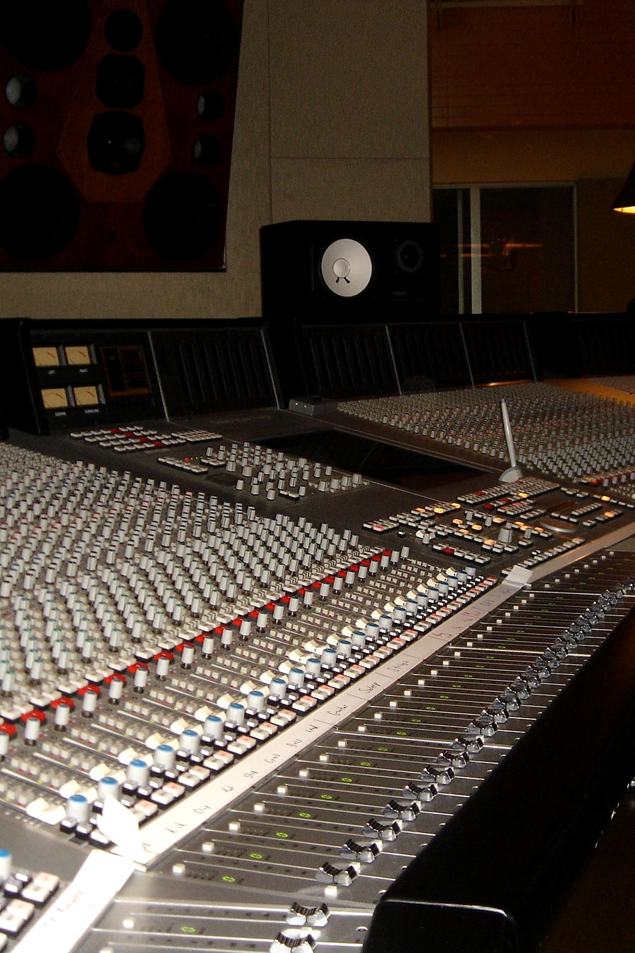 A large mixing console seen in perspective, with a window and a white-cone monitor beyond it.