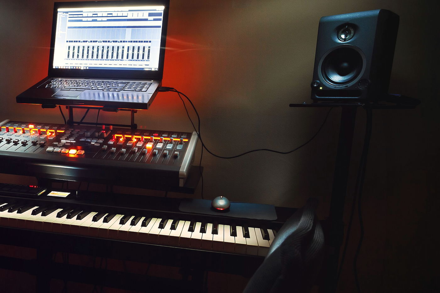 A laptop on a stand above a lit orange controller and a keyboard, with a black monitor on a wall arm against a red-lit wall.