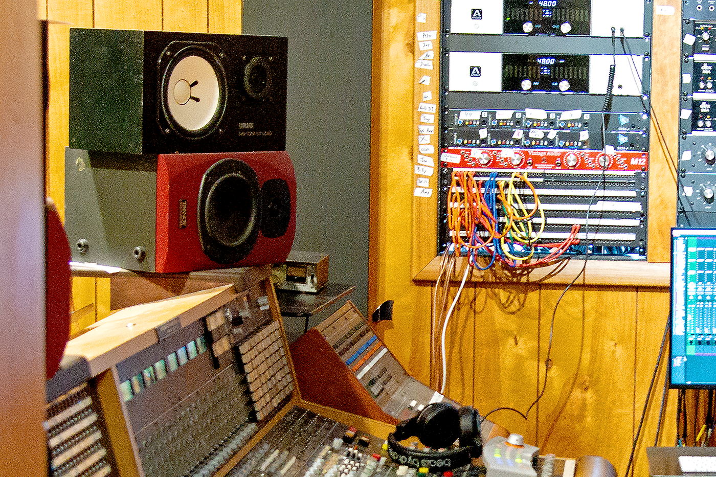 A white-cone near-field monitor standing on a red speaker, beside wood panelling, a rack of orange patch cables and a pair of headphones.