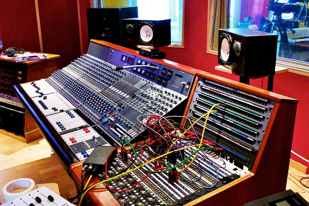 A vintage mixing console in a red-walled room, with colourful patch cables, three monitors on the meter bridge and a window behind.