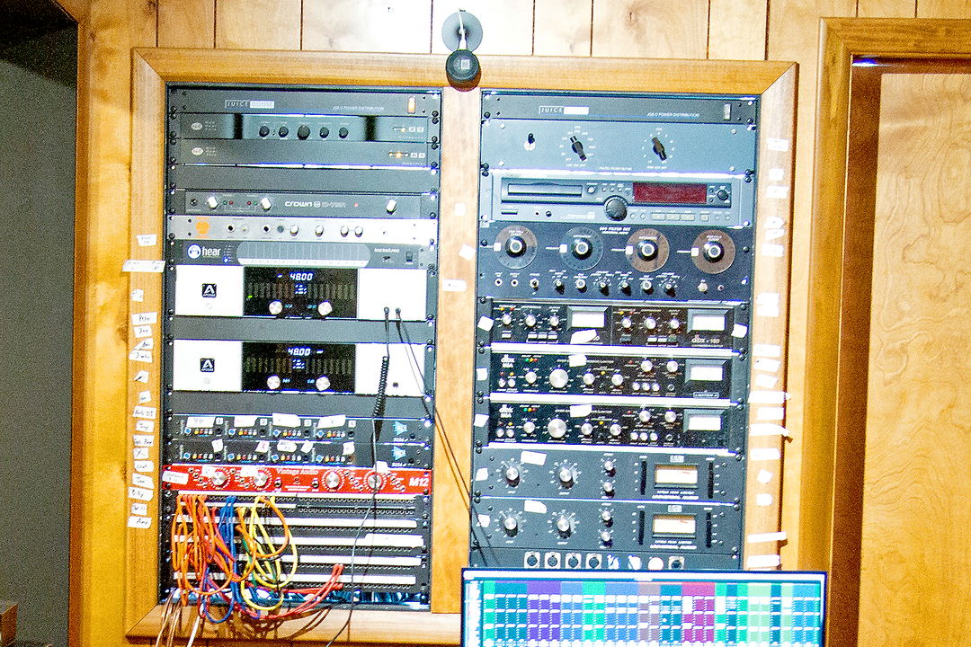 Two wood-framed equipment racks holding power amplifiers, compressors and a red patch bay with cables plugged in.