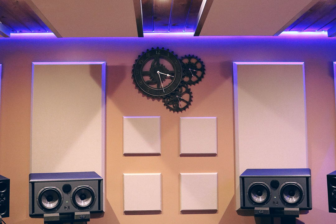 A cream-panelled studio wall with a gear-shaped clock under a purple-lit ceiling, above two large twin-woofer monitors.