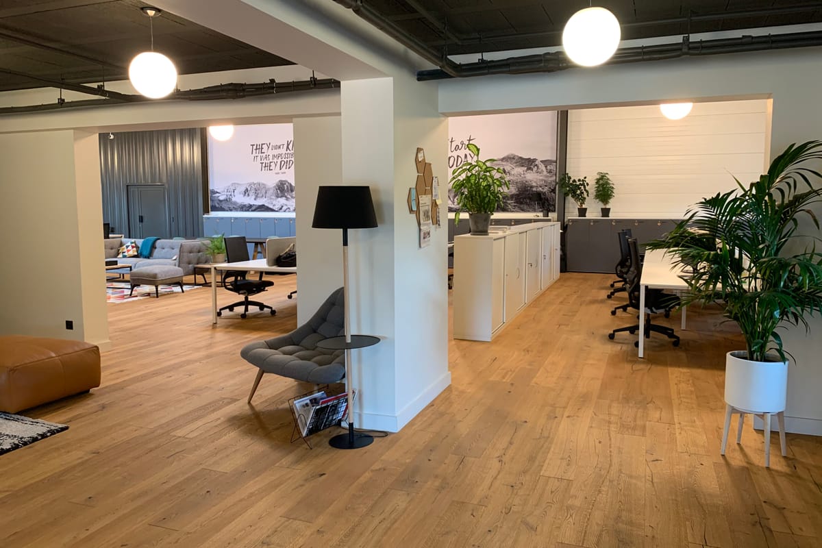 A coworking lounge with a sofa, low tables, desk partitions and plants on a pale wood floor.